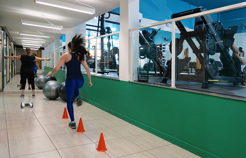 Foto de um corredor espaçoso da parte inferior da academia. Na imagem, uma figura feminina com roupas de ginástica corre pulando cones, enquanto outra figura feminina em roupas de ginástica se exercita em frente a um grande espelho no final do corredor.