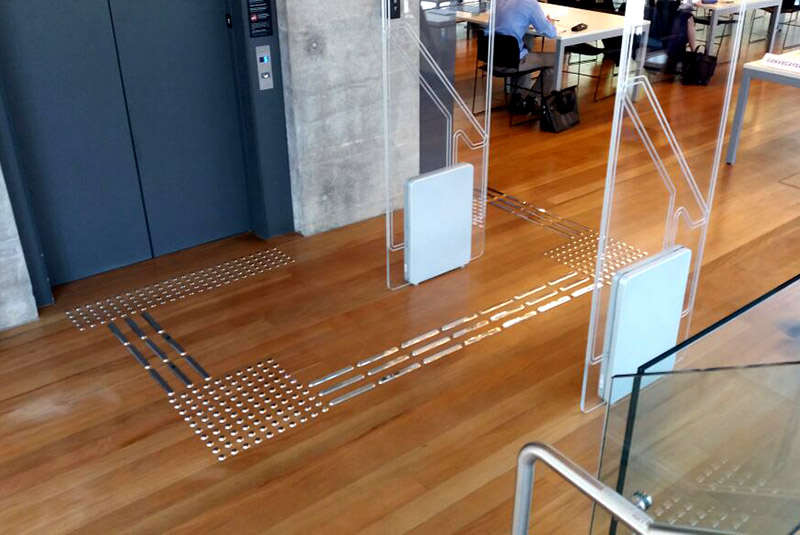 A imagem é uma foto aproximada do piso de alerta e direcional metálico sobre piso de madeira na frente do elevador.