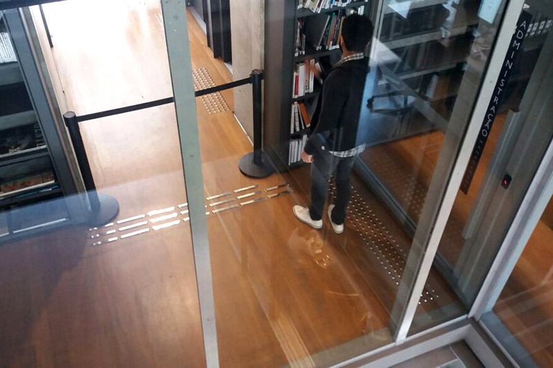 A imagem é uma foto vista do alto, mostrando uma pessoa procurando um livro numa prateleira. É possível ver o piso alerta e o piso direcional metálico sobre o piso de madeira do ambiente.