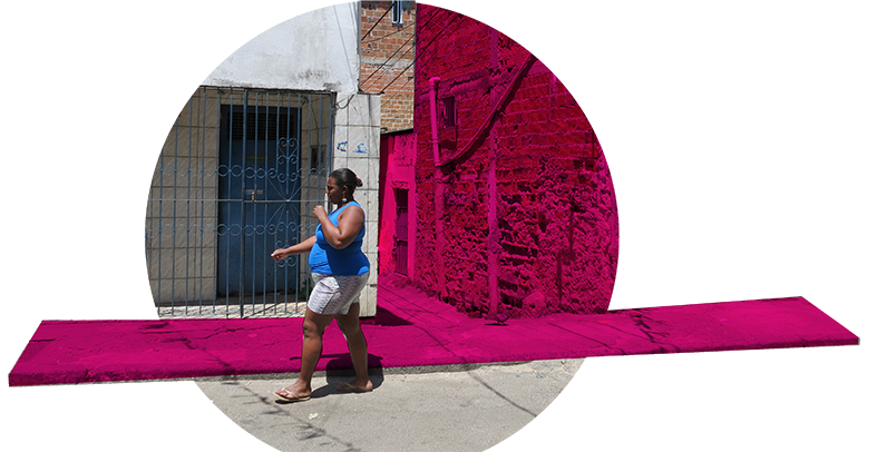 A imagem é uma montagem em cima de uma foto real. Temos uma mulher, pedestre, andando fora da calçada, que é estreita. Em rosa, destacamos o espaço da calçada e da viela ao fundo, mostrando o espaço público reduzido e de má qualidade reservado ao pedestre.
