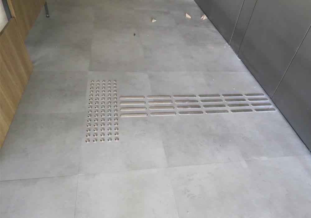 Foto do piso tátil já colocado. A imagem mostra um trecho de piso tátil de alerta e um trecho de piso de piso direcional, encaminhando o usuário ao balcão de informações.