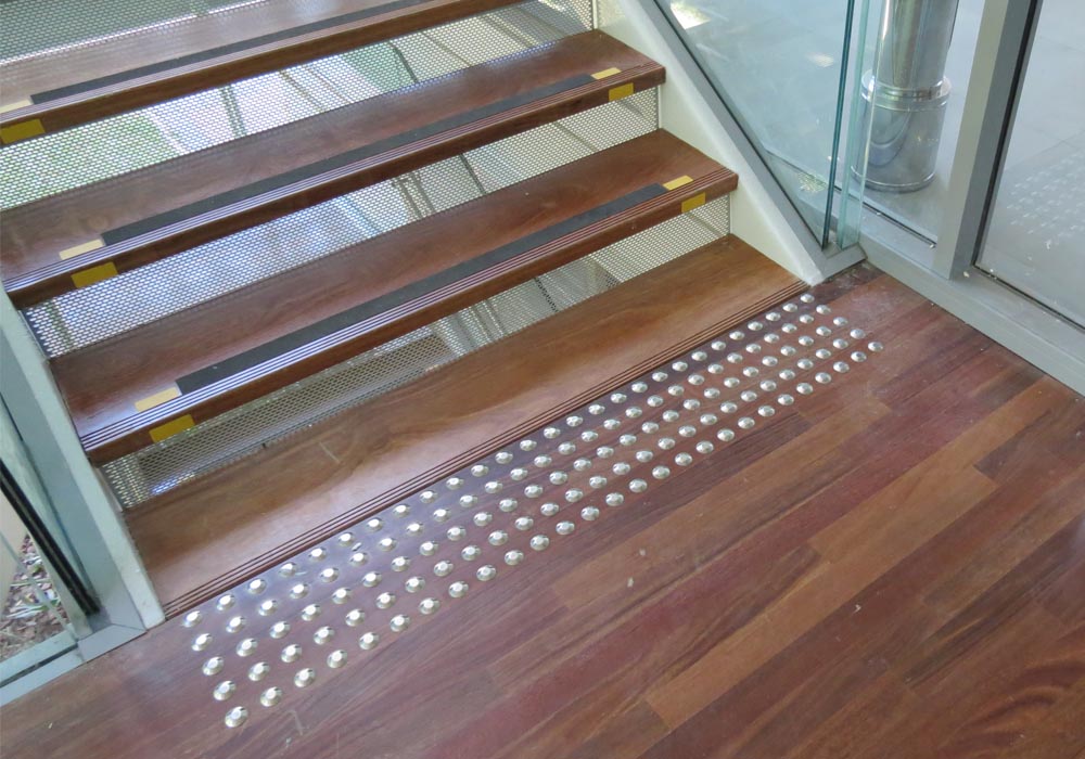 Foto de um trecho da escada, que mostra os três degraus inferiores sinalizados na lateral por fitas amarelas e com o piso tátil de alerta, em cor cinza claro. O piso é de madeira avermelhada.