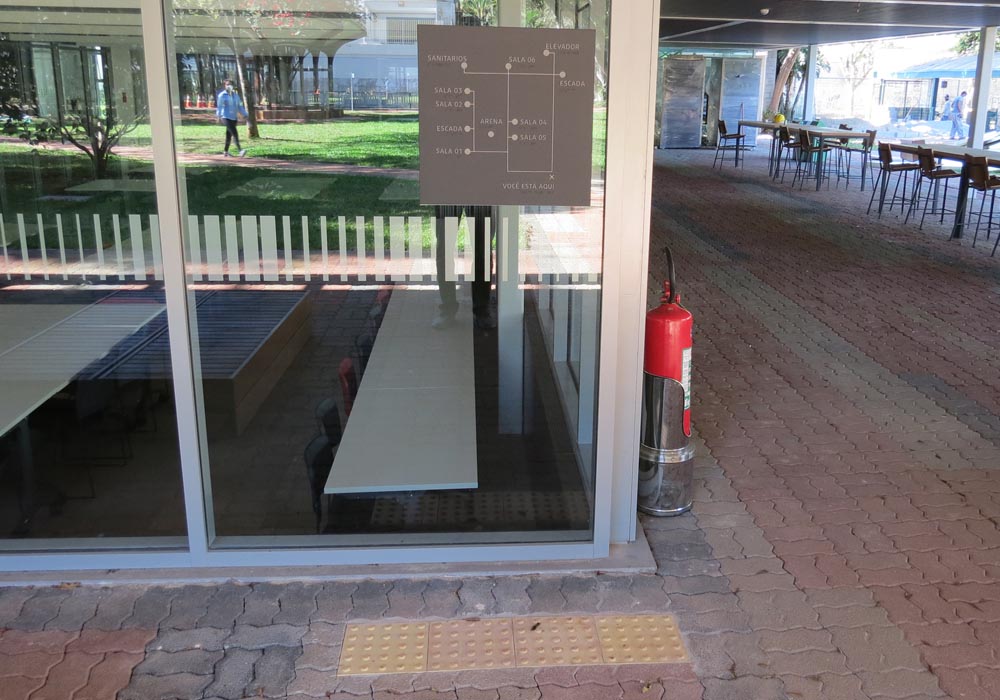 Foto do mapa tátil afixado em parede de vidro, com sinalização de alerta no piso. O mapa está em área externa da biblioteca, em local iluminado e há um reflexo de um gramado no vidro.